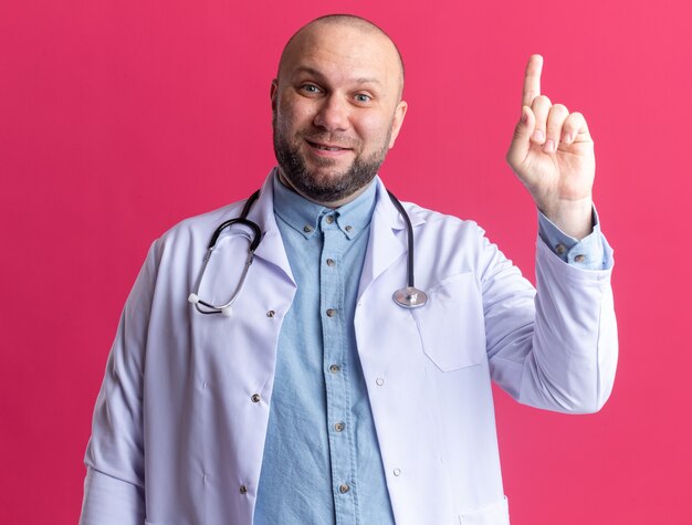 Beeindruckter männlicher Arzt mittleren Alters, der ein medizinisches Gewand und ein Stethoskop trägt, das isoliert auf einer rosa Wand nach oben zeigt