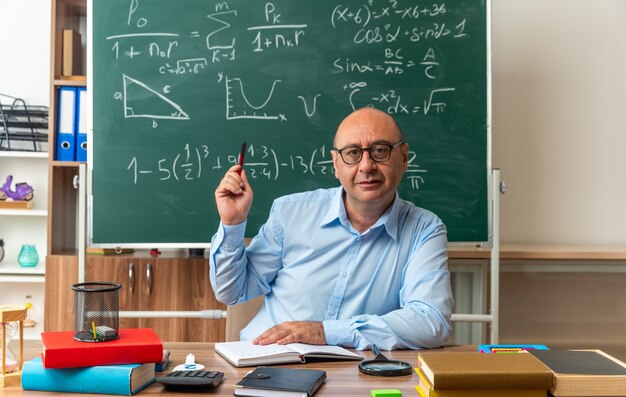 Beeindruckter männlicher Lehrer mittleren Alters mit Brille sitzt am Tisch mit Schulmaterial, das einen Stift im Klassenzimmer hält