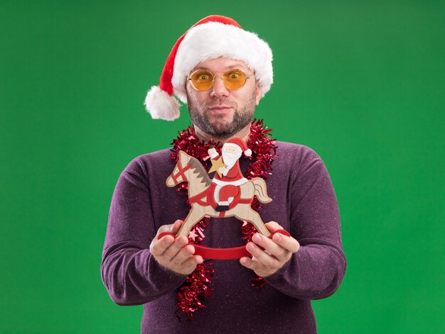 Beeindruckter Mann mittleren Alters mit Weihnachtsmütze und Lametta-Girlande um den Hals mit Brille, die den Weihnachtsmann auf einer Schaukelpferdefigur ausstreckt, die auf einer grünen Wand isoliert ist