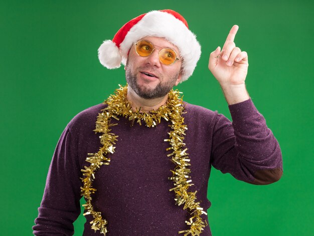 Beeindruckter Mann mittleren Alters mit Weihnachtsmütze und Lametta-Girlande um den Hals mit Brille, die die Seite betrachtet, die oben auf grünem Hintergrund zeigt