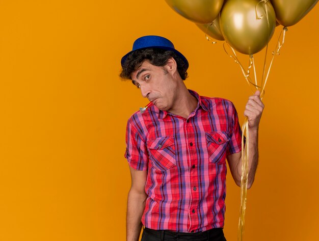 Beeindruckter Party-Mann mittleren Alters, der Partyhut hält, der Luftballons hält, die vorne mit Partygebläse im Mund lokalisiert auf orange Wand betrachten