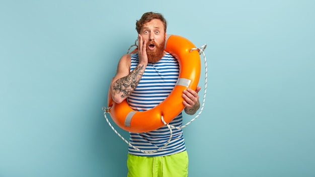 Beeindruckter rothaariger Mann hat einen nervösen Gesichtsausdruck, schwimmt zum ersten Mal im Meer und benutzt eine Ringboje