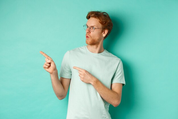 Beeindruckter rothaariger mann in brille und t-shirt, der mit den fingern zeigt und nach links auf das promo-angebot schaut, glücklich anstarrt und über türkisfarbenem hintergrund steht.