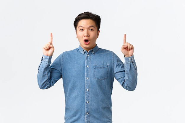 Beeindruckter und überraschter asiatischer Mann in blauem Hemd, der große Neuigkeiten diskutiert, mit den Fingern erstaunt nach oben zeigt, erstaunt in die Kamera schaut, interessante Verbindung gefunden, mit Menschen geteilt, weißer Hintergrund.