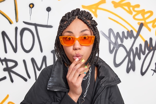 Beeindrucktes Hipster-Teenager-Mädchen mit Dreadlocks sieht sprachlos aus, wenn die Kamera die Hand auf den gefalteten Lippen hält. Sie trägt eine trendige orangefarbene Sonnenbrille und eine schwarze Jacke posiert an der Graffiti-Wand