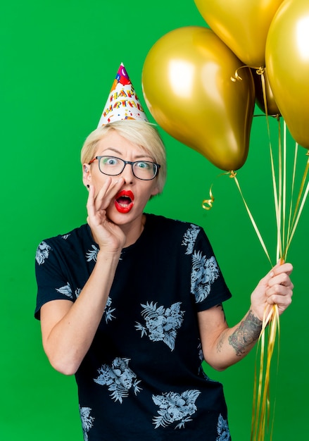 Beeindrucktes junges blondes Partygirl, das Brille und Geburtstagskappe hält, die Ballons hält, die Kamera betrachten und lokalisiert auf grünem Hintergrund flüstern