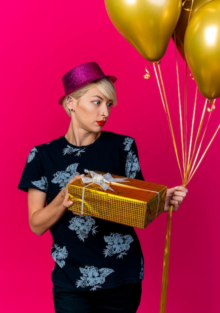 Beeindrucktes junges blondes Partygirl, das Partyhut hält, der Geschenkbox und Luftballons hält, die Seite lokal auf purpurrotem Hintergrund betrachten