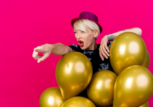 Kostenloses Foto beeindrucktes junges blondes partygirl, das partyhut trägt, der hinter luftballons steht und auf seite lokalisiert auf purpurrotem hintergrund mit kopienraum steht