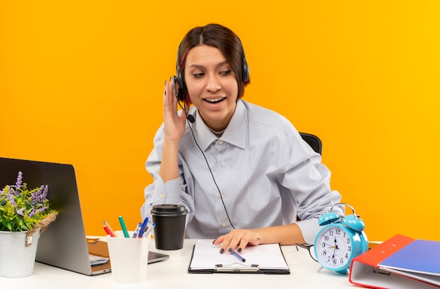 Kostenloses Foto beeindrucktes junges callcenter-mädchen, das ein headset trägt, das am schreibtisch sitzt und die hand auf das headset setzt, das die seite lokalisiert auf orange betrachtet