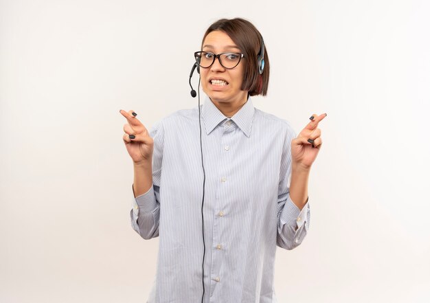 Beeindrucktes junges Callcenter-Mädchen, das eine Brille und ein Headset trägt, die ihre Finger kreuzen, lokalisiert auf Weiß mit Kopienraum