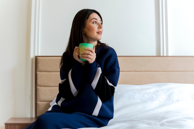 Beeindrucktes junges hübsches kaukasisches Mädchen, das auf Bett im Schlafzimmer sitzt Tasse hält und Seite betrachtet