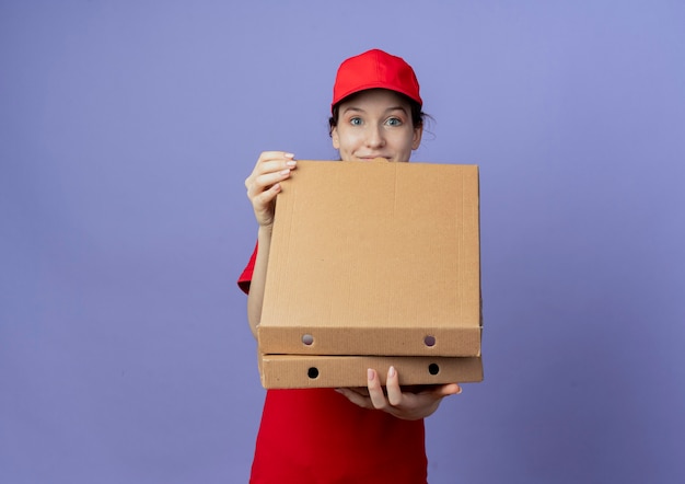 Beeindrucktes junges hübsches Liefermädchen, das rote Uniform und Kappe hält und Pizzapakete auf lila Hintergrund mit Kopienraum isoliert