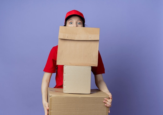 Beeindrucktes junges hübsches Liefermädchen in roter Uniform und Mütze, das Kartons und Papierpaket hält und von hinten Papierpaket schaut
