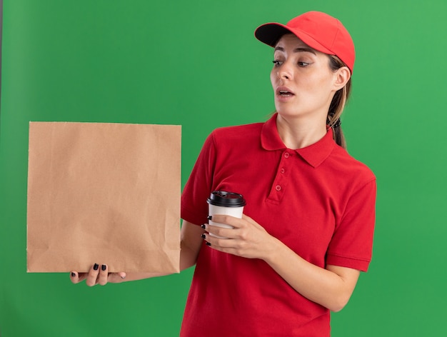 Beeindrucktes junges hübsches Liefermädchen in Uniform hält Pappbecher und schaut auf Papierpaket auf Grün