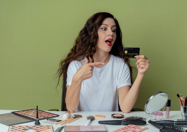 Beeindrucktes junges hübsches Mädchen, das am Make-up-Tisch mit Make-up-Tools sitzt und auf die Kreditkarte auf dem olivgrünen Raum schaut und auf sie zeigt