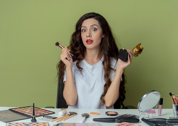 Beeindrucktes junges hübsches Mädchen, das am Make-up-Tisch mit Make-up-Werkzeugen hält, die Puderpinsel und Siegerbecher lokalisiert auf olivgrünem Hintergrund halten
