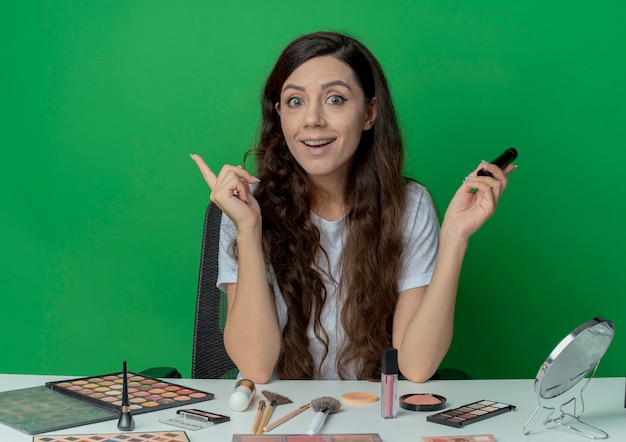 Beeindrucktes junges hübsches Mädchen, das am Make-up-Tisch mit Make-up-Werkzeugen hält, die Wimperntusche halten und Finger lokalisiert auf grünem Hintergrund halten