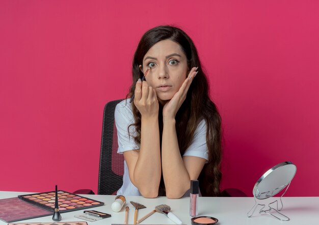 Beeindrucktes junges hübsches Mädchen, das am Make-up-Tisch mit Make-up-Werkzeugen sitzt, die Hand auf Gesicht setzen und Lidschatten mit Pinsel lokalisiert auf purpurrotem Hintergrund anwenden