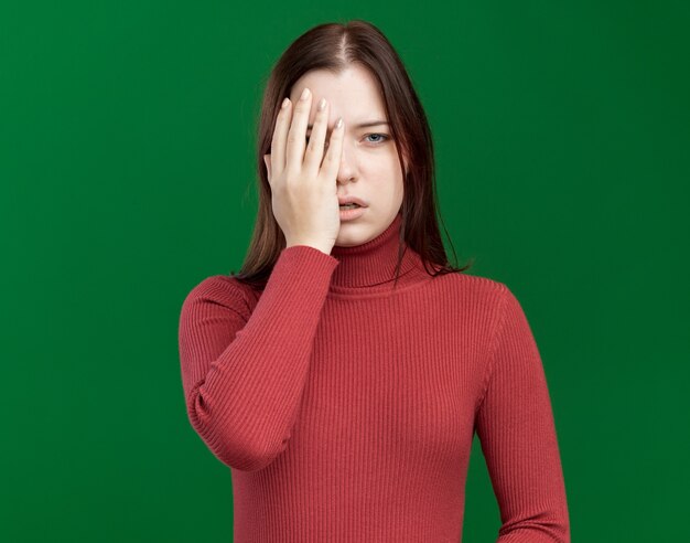 Beeindrucktes junges hübsches Mädchen, das die Hälfte des Gesichts mit der Hand bedeckt, isoliert auf grüner Wand mit Kopierraum