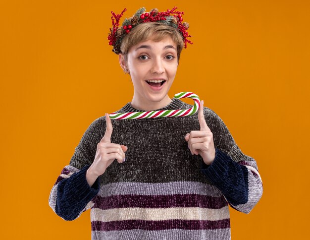 Beeindrucktes junges hübsches Mädchen, das Weihnachtskopfkranz hält, der weihnachtliches süßes Rohr an der Kamera lokalisiert auf orange Hintergrund hält