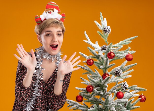 Beeindrucktes junges hübsches Mädchen, das Weihnachtsmann-Stirnband und Lametta-Girlande um den Hals trägt, der nahe verziertem Weihnachtsbaum steht, der leere Hände lokalisiert auf orange Wand zeigt