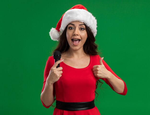 Beeindrucktes junges hübsches Mädchen, das Weihnachtsmütze hält Mikrofon, das Kamera betrachtet Daumen oben auf grünem Hintergrund zeigt