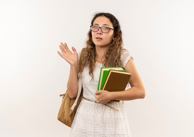 Beeindrucktes junges hübsches Schulmädchen, das Brille und Rückentasche hält Bücher hält, die leere Hand zeigen, die lokal auf Weiß schaut