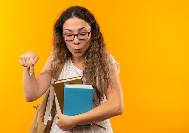 Beeindrucktes junges hübsches Schulmädchen, das Brille und Rückentasche hält Bücher hält, die lokal auf Gelb schauen und zeigen