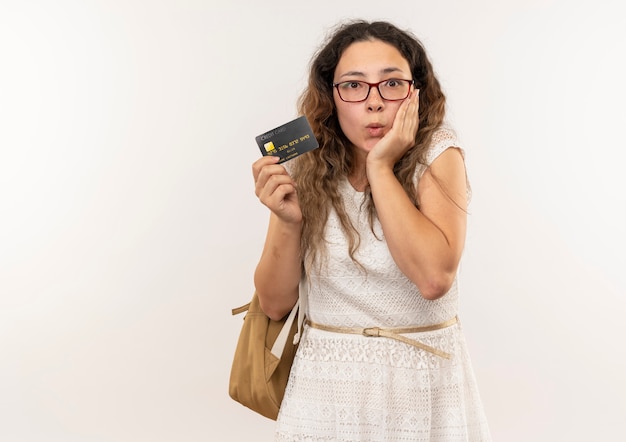 Beeindrucktes junges hübsches Schulmädchen, das Brille und Rückentasche hält, die Kreditkarte mit Hand auf Gesicht lokalisiert auf Weiß hält