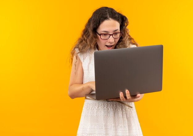 Beeindrucktes junges hübsches Schulmädchen, das Brille und Rückentasche unter Verwendung des auf Gelb isolierten Laptops trägt