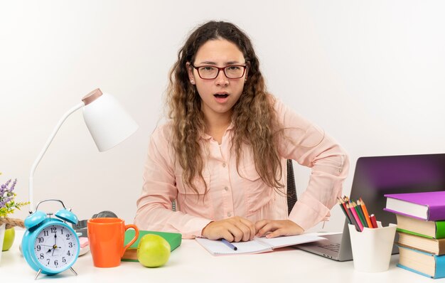 Beeindrucktes junges hübsches Schulmädchen, das eine Brille trägt, die am Schreibtisch mit Schulwerkzeugen sitzt, die ihre Hausaufgaben lokal auf Weiß tun