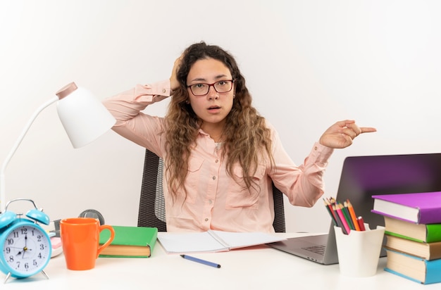 Kostenloses Foto beeindrucktes junges hübsches schulmädchen, das eine brille trägt, die am schreibtisch mit schulwerkzeugen sitzt, die ihre hausaufgaben machen, hand auf kopf setzen und auf seite zeigen, die auf weiß isoliert ist