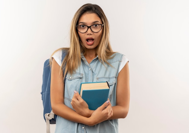 Beeindrucktes junges hübsches Studentenmädchen, das Brille und Rückentasche hält Buch lokalisiert auf Weiß trägt