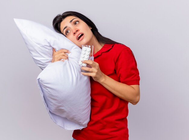 Beeindrucktes junges kaukasisches krankes Mädchen, das Kissen umarmt, das den Kopf darauf setzt, der gerade mit Packung der Tabletten und Glas Wasser in der Hand lokalisiert auf weißem Hintergrund mit Kopienraum schaut