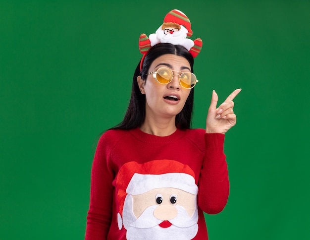 Beeindrucktes junges kaukasisches Mädchen, das Weihnachtsmann-Stirnband und Pullover mit Brille trägt, die lokal auf grünem Hintergrund suchen und zeigen