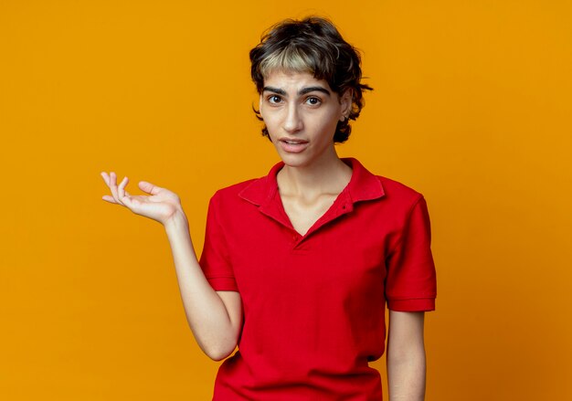 Beeindrucktes junges kaukasisches Mädchen mit Pixie-Haarschnitt, der leere Hand lokalisiert auf orange Hintergrund mit Kopienraum zeigt
