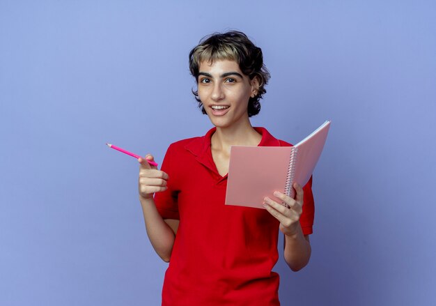 Beeindrucktes junges kaukasisches Mädchen mit Pixie-Haarschnitt, der Notizblock und Stift hält und auf Kamera mit Finger lokalisiert auf lila Hintergrund mit Kopienraum zeigt
