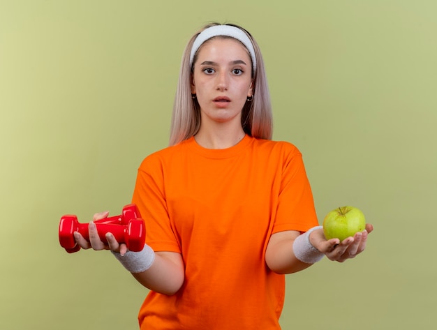 Beeindrucktes junges kaukasisches sportliches Mädchen mit Hosenträgern, das Stirnband und Armbänder trägt, hält Hanteln und Apfel