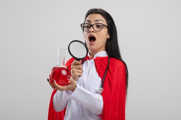 Beeindrucktes junges kaukasisches Superheldenmädchen, das Brille und Stethoskop hält und chemischen Kolben mit Lupe hält, lokalisiert auf weißem Hintergrund mit Kopienraum