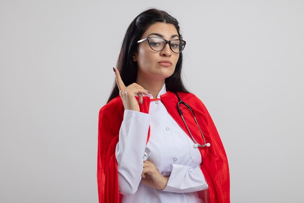 Beeindrucktes junges kaukasisches Superheldenmädchen, das Brille und Stethoskop trägt und nach unten zeigt, lokalisiert auf weißem Hintergrund mit Kopienraum
