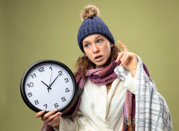 Kostenloses Foto beeindrucktes junges krankes mädchen, das seitliches tragen des weißen gewandes und der wintermütze mit schal hält wanduhr in plaidspitzen an oben auf olivgrün lokalisiert