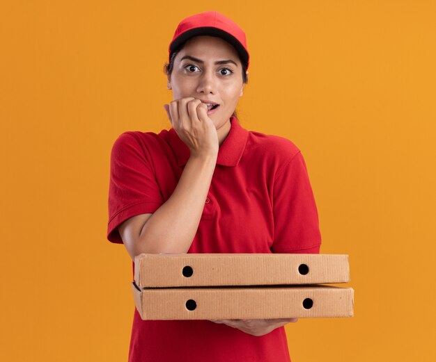 Beeindrucktes junges Liefermädchen, das Uniform und Kappe hält, die Pizzakästen hält, beißt Nägel lokalisiert auf orange Wand