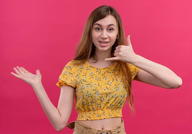 Beeindrucktes junges Mädchen, das Anrufgeste tut und leere Hand auf lokalisiertem rosa Raum zeigt