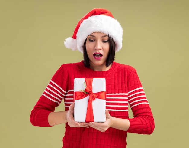 Beeindrucktes junges Mädchen, das Weihnachtsmütze hält und Geschenkpaket lokalisiert auf olivgrünem Hintergrund hält