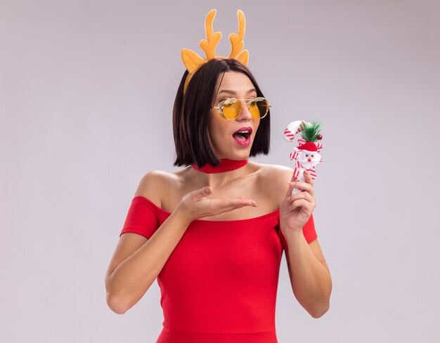 Beeindrucktes junges Mädchen mit Rentiergeweih-Stirnband und Brille mit Blick auf das Zeigen auf Weihnachtszuckerstangenverzierung isoliert auf weißem Hintergrund