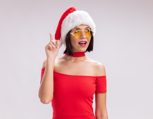 Beeindrucktes junges Mädchen mit Weihnachtsmütze und Brille, das auf die Seite schaut, die nach oben zeigt, isoliert auf weißem Hintergrund mit Kopierraum