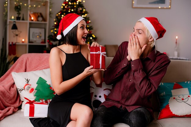 Beeindrucktes junges Paar zu Hause zur Weihnachtszeit mit Weihnachtsmütze auf Sofa im Wohnzimmer Mädchen geben Geschenkpaket zu ihrem Freund, der ihn ansieht, er schaut auf Geschenk, das Hände auf Mund hält