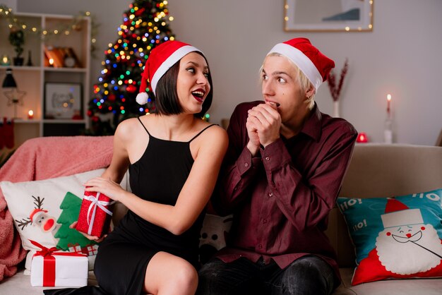 Beeindrucktes junges Paar zu Hause zur Weihnachtszeit mit Weihnachtsmütze auf Sofa im Wohnzimmer sitzend Geschenke