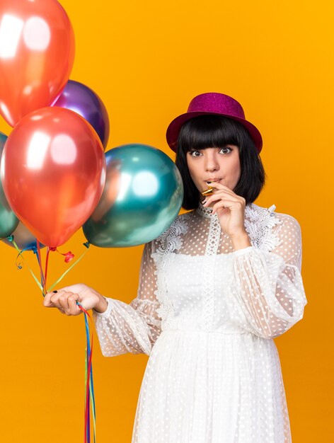 Beeindrucktes junges Partymädchen mit Partyhut, der Luftballons hält, die Partyhorn im Mund halten, isoliert auf oranger Wand