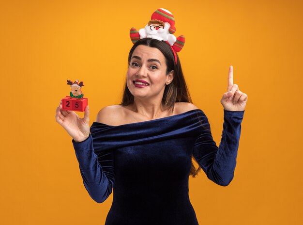 Beeindrucktes junges schönes Mädchen, das blaues Kleid und Weihnachtshaarbügel hält, der Spielzeugpunkte an oben lokalisiert auf orangefarbenem Hintergrund hält